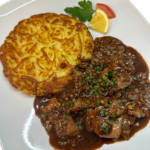 Plate with crispy potato rosti and sliced beef in brown mushroom gravy, garnished with a lemon wedge, tomato slice and parsley leaves. Kalbs Leber mit knusprigen Schweizer Rösti
